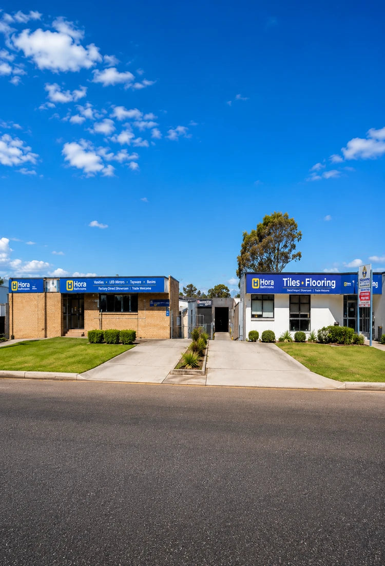 Two small commercial buildings with blue signs reading 'HORA BATHROOMS' under a bright blue sky with scattered clouds, set behind a paved road and grassy lawns.